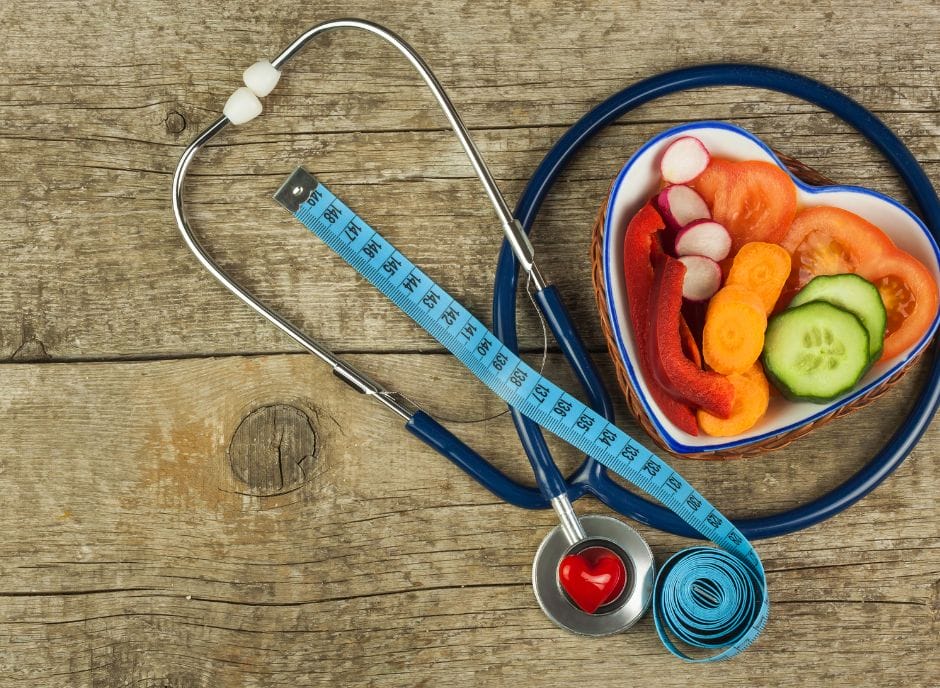 A heart shaped bowl of vegetables and a stethoscope on a wooden table.