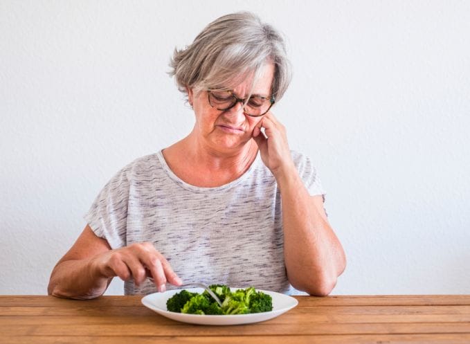 A woman sitting at a table with a plate of broccoli, following a renal diet.