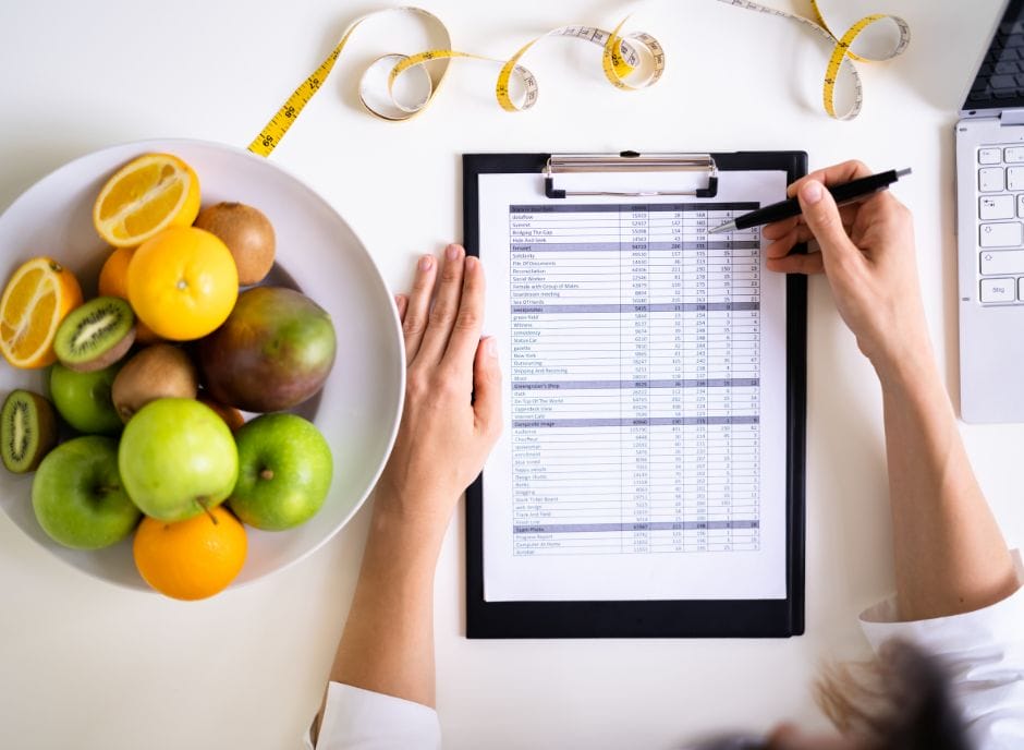 A woman writing a diet plan on a clipboard in front of a bowl of fruit.