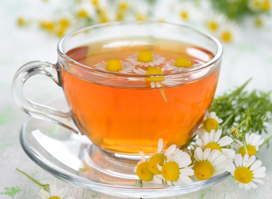 A clear glass cup of chamomile tea with fresh chamomile flowers on the side.