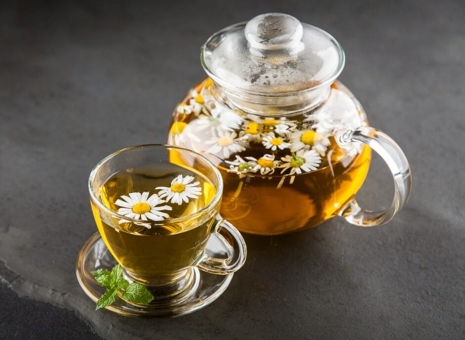 A glass teapot with chamomile tea and fresh flowers inside, alongside a matching cup filled with tea on a dark surface.