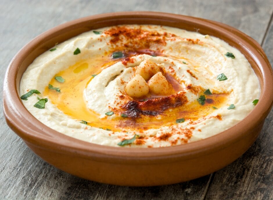 A bowl of creamy hummus garnished with chickpeas, olive oil, paprika, and chopped parsley on a wooden table.