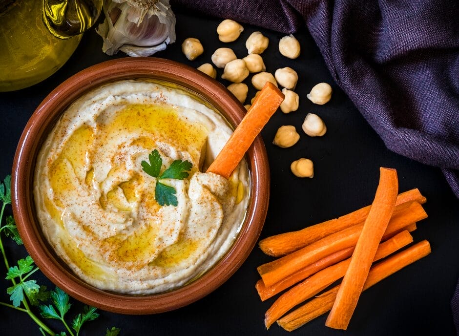 A bowl of hummus garnished with parsley and paprika, surrounded by carrot sticks, chickpeas, and romaine lettuce on a dark background.