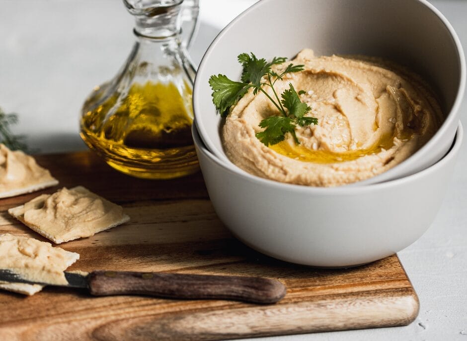 A bowl of hummus topped with olive oil and parsley on a wooden board, with a jar of olive oil beside it and a romaine lettuce leaf dipped in hummus.