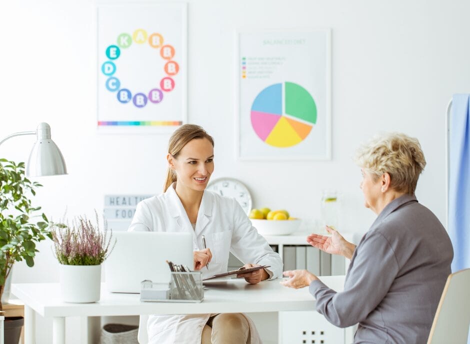 A female renal dietitian in a white coat discussing nutrition with an elderly female patient in a bright office, charts on the wall.