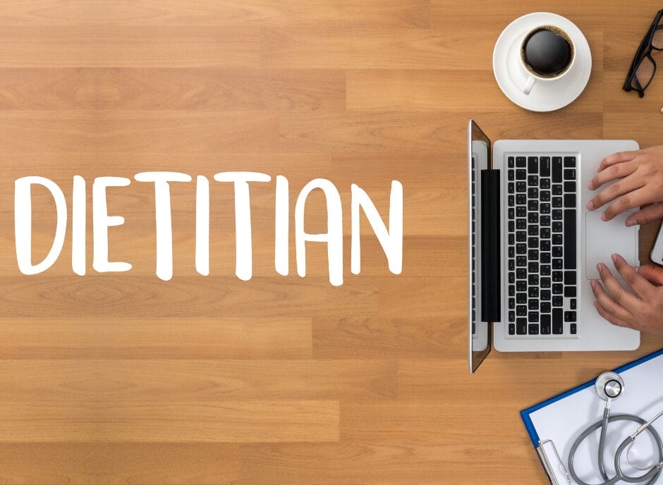 Overhead view of a workspace with the word "dietitian" on it, featuring hands typing on a laptop, a stethoscope, notepad, and a cup of coffee on a wooden desk.