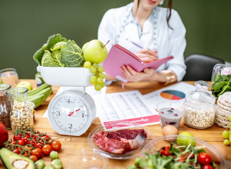 A dietitian sits at a table surrounded by a variety of healthy foods and a scale, writing in a notebook.