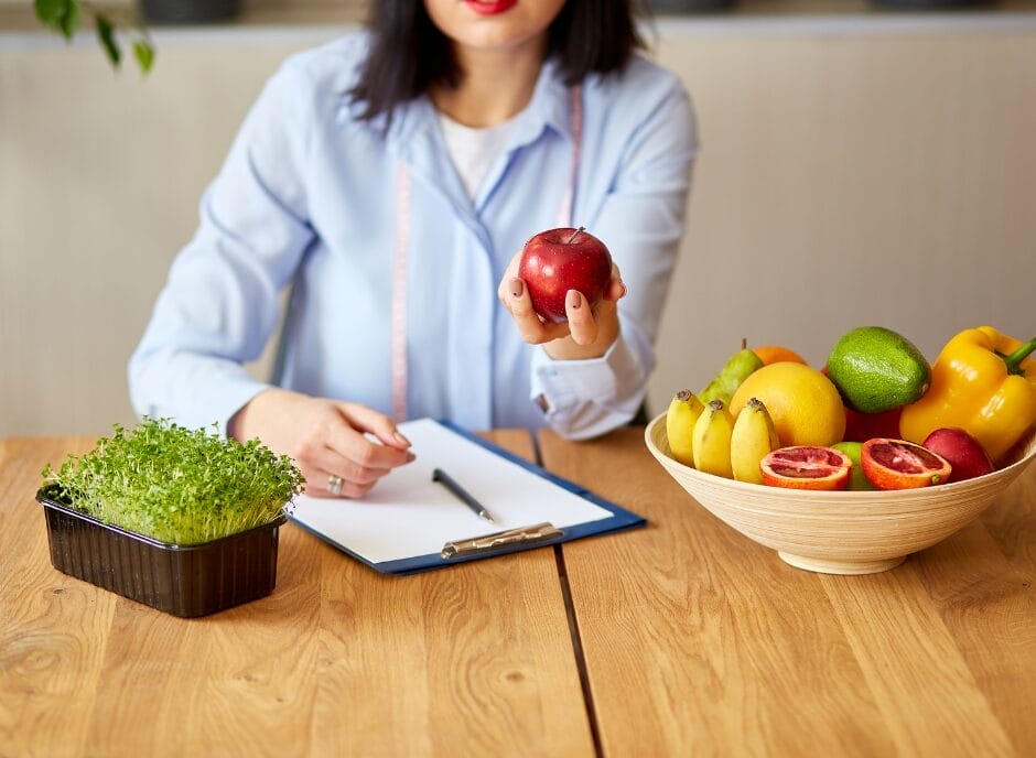 A woman sits at a desk holding a red apple, with a notepad beside her and a bowl of assorted fruits and a tray of sprouts on the table.