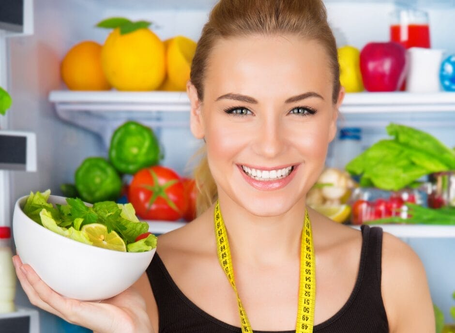 A young woman with a measuring tape around her neck smiles while holding a bowl of salad, standing in front of an open refrigerator stocked with fresh produce and beverages.
